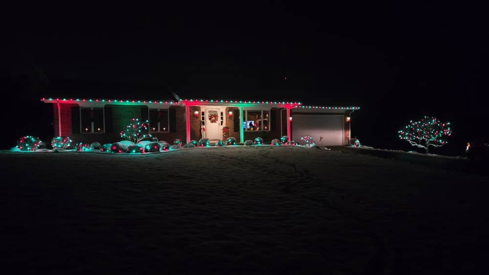 Ranch home with red and green roofline lights and lit shrubs in snow